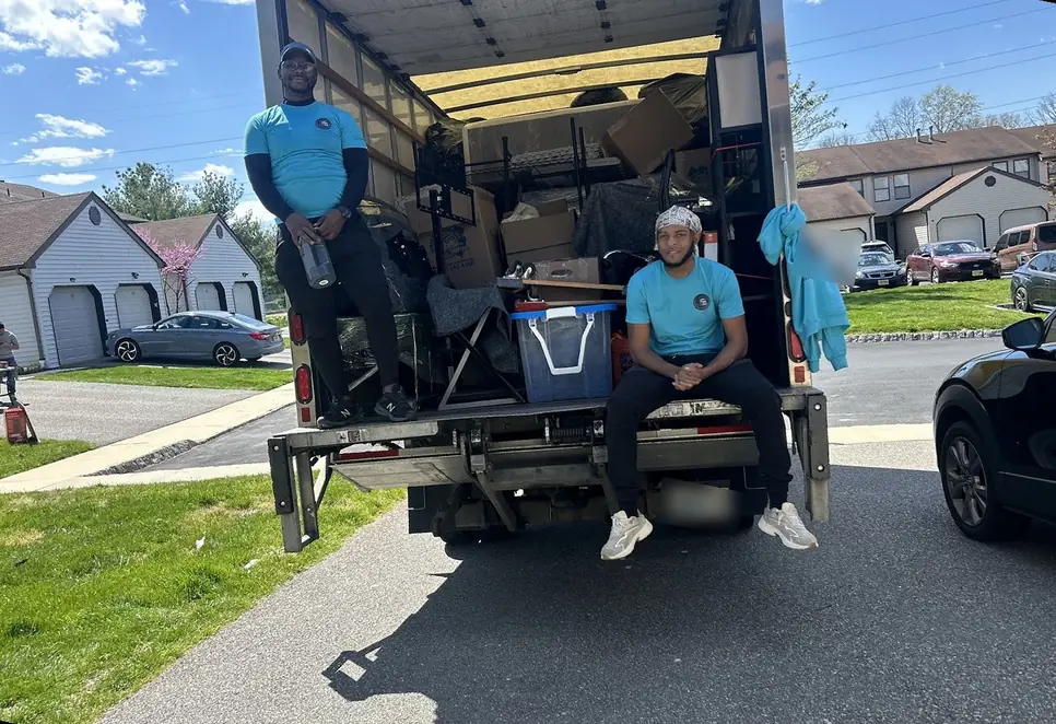 Two B&M movers in teal polos sitting on the liftgate of a loaded truck on a townhouse street